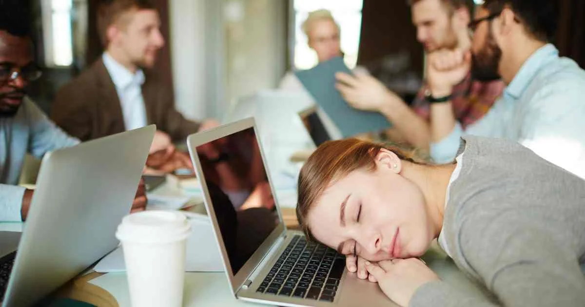  A tired woman resting her head on a laptop in a busy office setting while coworkers discuss in the background.